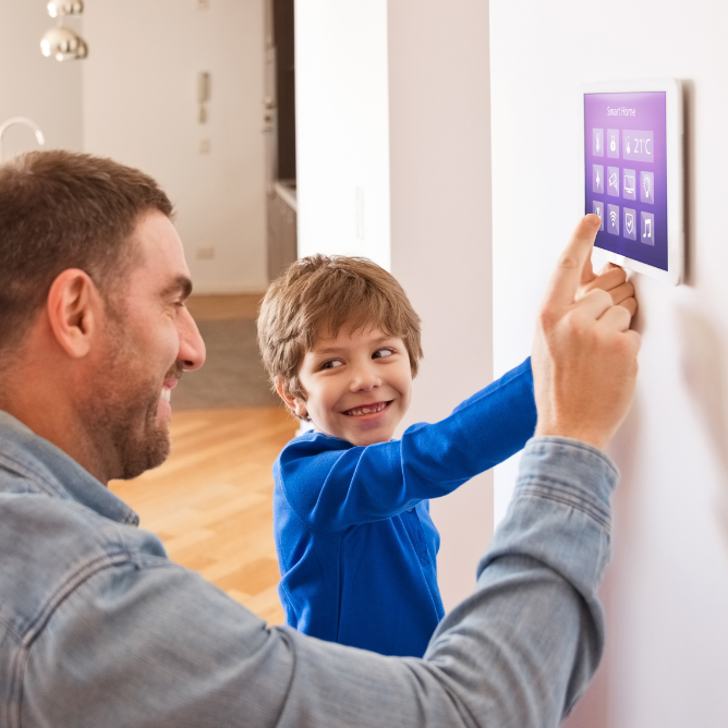 A man and a young boy are using a smart home system that is mounted on the wall. They are both smiling.