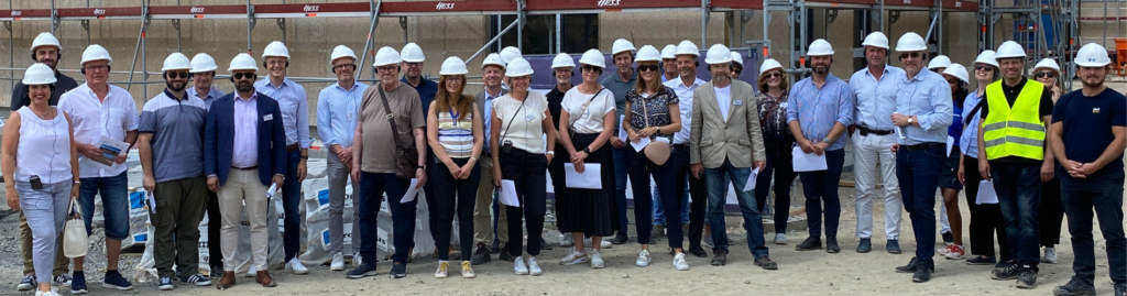 Group of people in white hard hats standing smiling in front of construction site. Public and social housing network.