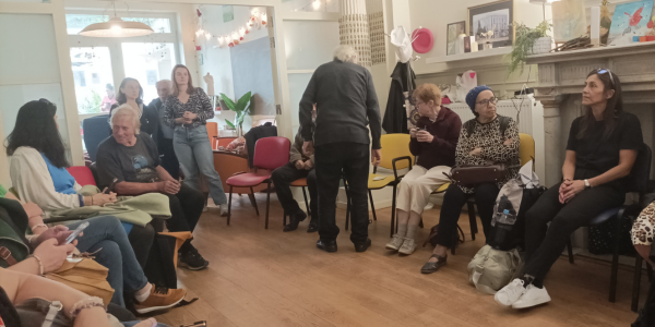 Inside Biloba Huis. A group of people sit in a circle on chairs chatting between each other. The background is cheerful with decorations.