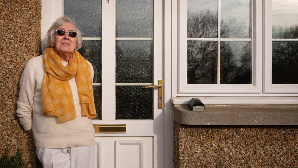 An older woman stands leaning against her front door. She is weating white trousers, a cream jumper and a bright yellow scarf. She also wears sunglasses.
