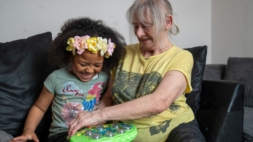 An older woman wearing a bright yellow t-shirt plays with a young girl wearing a flower crown while seated on a sofa. Both are smiling.