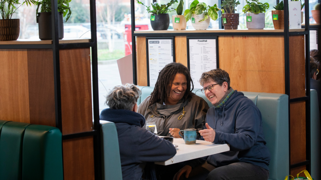 Three people sit at a table in a cafe laughing. They are drinking hot drinks and look to be aged 50+.