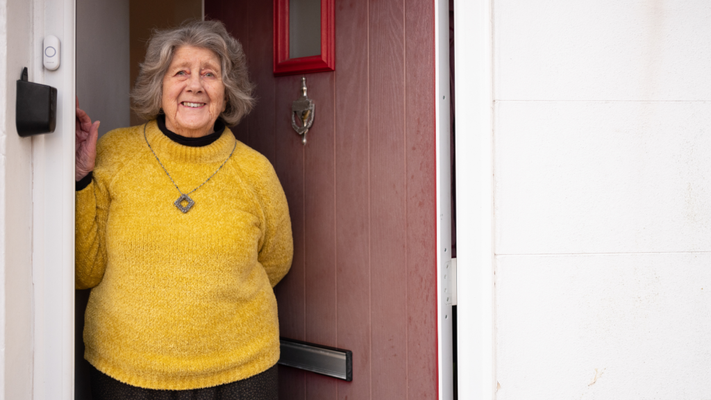 An older woman stands in the doorway of her home smiling. She wears a bright yellow jumper and a silver necklace in a diamond shape. The door is brown-red with a bright red window.